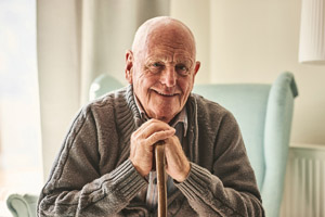 Elderly man sitting and holding a walking stick, smiles at the camera